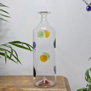 May include: A clear glass bottle with a long neck and a flat base. The bottle is decorated with colorful polka dots in yellow, green, and blue. The bottle is displayed on a wooden surface, with green plants in the background.