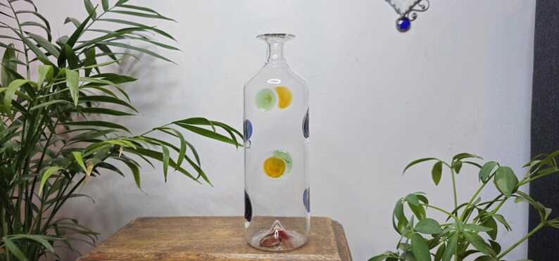 May include: A clear glass bottle with a long neck and a flat base. The bottle is decorated with colorful polka dots in yellow, green, and blue. The bottle is displayed on a wooden surface, with green plants in the background.