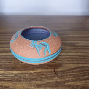 May include: A small, round, terracotta-colored ceramic bowl with a speckled finish. The exterior features a turquoise blue Kokopelli figure and two horizontal turquoise blue bands encircling the lower portion. The interior is a solid light purple.