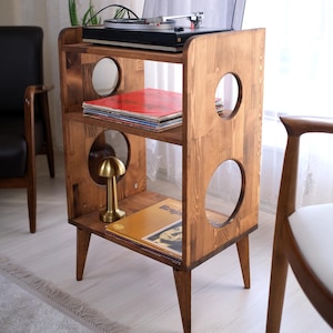 May include: A wooden record player stand with three shelves and circular cutouts. The stand holds a record player, vinyl records, and a small gold lamp. The wood has a warm, natural finish, and the stand has angled legs.
