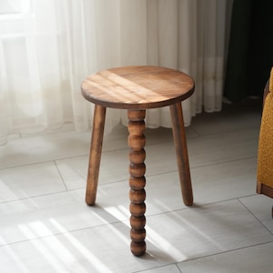 May include: A wooden stool with a round seat and three legs. The centre leg is made of a series of wooden beads.