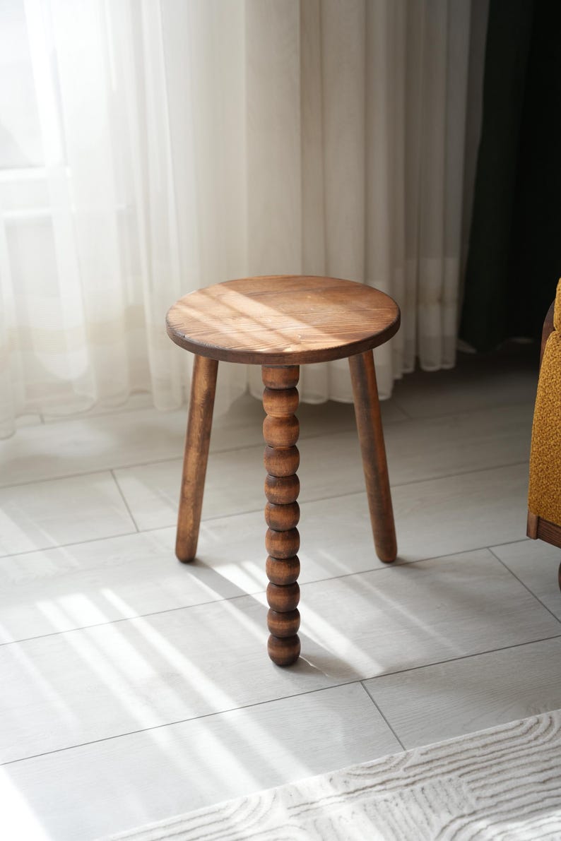 May include: A wooden stool with a round seat and three legs. The center leg is made of a series of wooden beads.