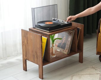 May include: A wooden record player stand with a built-in record storage shelf. The stand is stained a warm brown color and holds a black turntable with a clear dust cover. Several vinyl records are visible on the shelf.