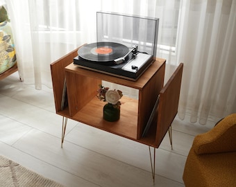 May include: A wooden record player stand with a black turntable playing a vinyl record. The stand has a lower shelf with a small vase of flowers and gold-colored legs. The stand is made of light brown wood.