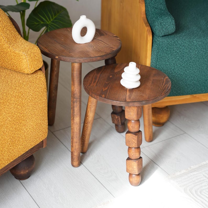 May include: Two wooden side tables with different leg designs. The table on the left has three straight legs and the table on the right has one leg with a stacked cube design. Both tables have a dark brown wood finish.