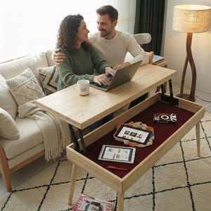 May include: A light wood lift-top coffee table with a lower storage compartment. The table has a rectangular top and four legs. The lower compartment is lined with a burgundy fabric and contains two tablets and a book. A couple is sitting at the table.