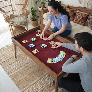 Op de afbeelding: Een houten speeltafel met een bordeauxrode viltlaag. Er zijn verschillende kleurrijke kaarthouders op de tafel geplaatst. Twee mensen spelen een kaartspel, met kaarten in hun handen. De tafel staat in een woonkamer.
