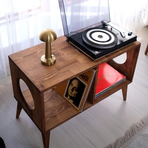 以下が含まれることがあります： 木製のレコードプレーヤー台。レコード用の棚付き。ミッドセンチュリーモダンデザインで、濃い茶色の仕上げと金色のキノコランプが付いています。黒いレコードプレーヤーが上にあります。長さ76cm。