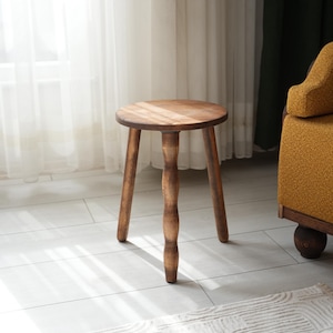 May include: A wooden stool with a round seat and three legs. The legs are turned and have a decorative design. The stool is brown and has a natural finish.