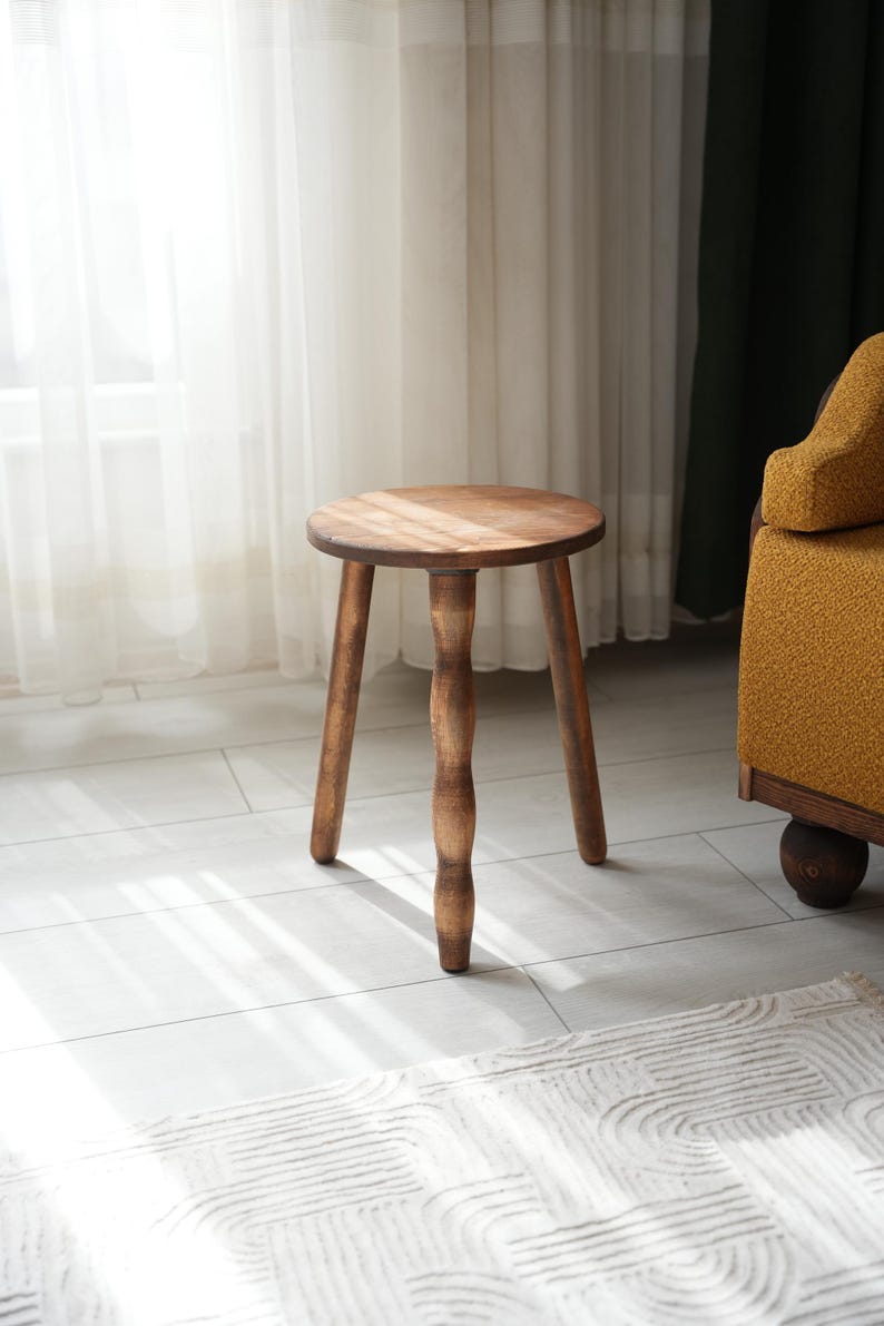 May include: A wooden stool with a round seat and three legs. The legs are turned and have a decorative design. The stool is brown and has a natural finish.