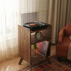 May include: A dark brown wooden record player stand with a record player on top. The stand has two shelves, one with a clock and the other with vinyl records. A small vase with a green plant is also on the shelf. The stand is on a patterned rug.