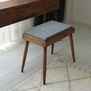 May include: A wooden stool with a gray upholstered seat. The stool has four angled, tapered legs and a rectangular frame. The wood has a warm, brown tone. The stool is placed on a light-colored rug.