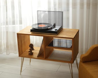 May include: A wooden record player stand with a black turntable and a record playing on top. The stand has a lower shelf for record storage and gold-colored hairpin legs. A small statue is on the lower shelf.