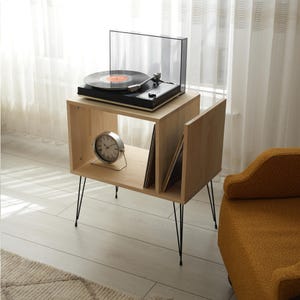 Puede incluir: Un soporte de tocadiscos de madera clara con patas de horquilla negras. Un tocadiscos negro con una cubierta transparente se encuentra en la parte superior. Un reloj plateado y discos de vinilo se guardan en los compartimentos abiertos. El soporte está junto a un sillón amarillo.