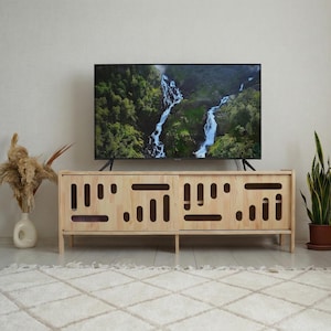 Pode incluir: Um móvel de TV de madeira clara com formato retangular e desenhos recortados. Uma TV de tela plana grande exibe uma cena de cachoeira. O móvel está sobre um tapete branco, e um vaso com plantas secas está à esquerda.