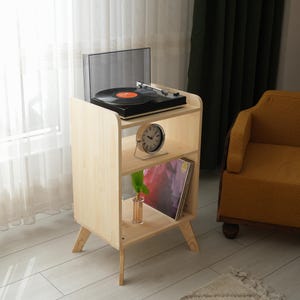 Puede incluir: Un soporte para tocadiscos de madera clara con un tocadiscos negro en la parte superior. El soporte tiene dos estantes, uno con un reloj y el otro con un álbum. El soporte está al lado de un sillón amarillo.