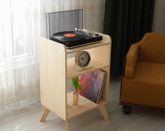 Puede incluir: Un soporte para tocadiscos de madera clara con un tocadiscos negro en la parte superior. El soporte tiene dos estantes, uno con un reloj y el otro con un álbum. El soporte está al lado de un sillón amarillo.