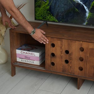 May include: A dark brown wooden TV stand with a sliding door featuring circular cutouts. The stand has a lower shelf with books that read "CHANEL", "Dior", and "KINFOLK". A flat-screen TV sits on top.