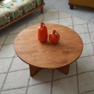 May include: A round wooden coffee table with a natural finish. Two orange vases with gold accents sit on the table. The table is approximately 36 inches in diameter and has a four-legged base. The table is placed on a patterned rug.