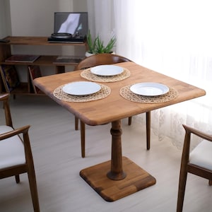 Può includere: Un tavolo da pranzo quadrato in legno con base a piedistallo, apparecchiato con tre piatti bianchi su tovagliette intrecciate. Sono visibili due sedie in legno con cuscini bianchi. Il tavolo misura circa 76 cm quadrati.