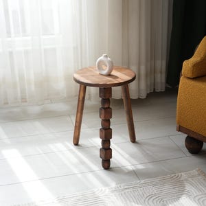 May include: A wooden side table with a round top and three legs. The table has a unique design with a stack of wooden blocks forming one of the legs. A white ceramic vase sits on the table top.