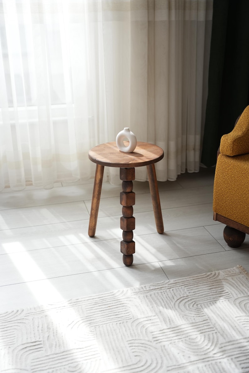 May include: A wooden side table with a round top and three legs. The table has a unique design with a stack of wooden blocks forming one of the legs. A white ceramic vase sits on the table top.