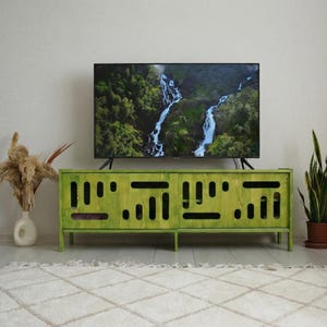 Könnte beinhalten: Ein limettengrüner TV-Schrank aus Holz mit rechteckiger Form und Aussparungsdesign. Der Schrank befindet sich unter einem großen Flachbildfernseher, der eine Wasserfallszene zeigt. Eine weiße Vase mit getrockneten Pflanzen und eine Topf-Schlangenpflanze befinden sich auf beiden Seiten.