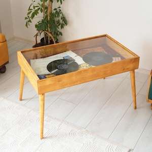 May include: A wooden coffee table with a glass top. The table has a light wood finish and four legs. There are two vinyl records inside the table.