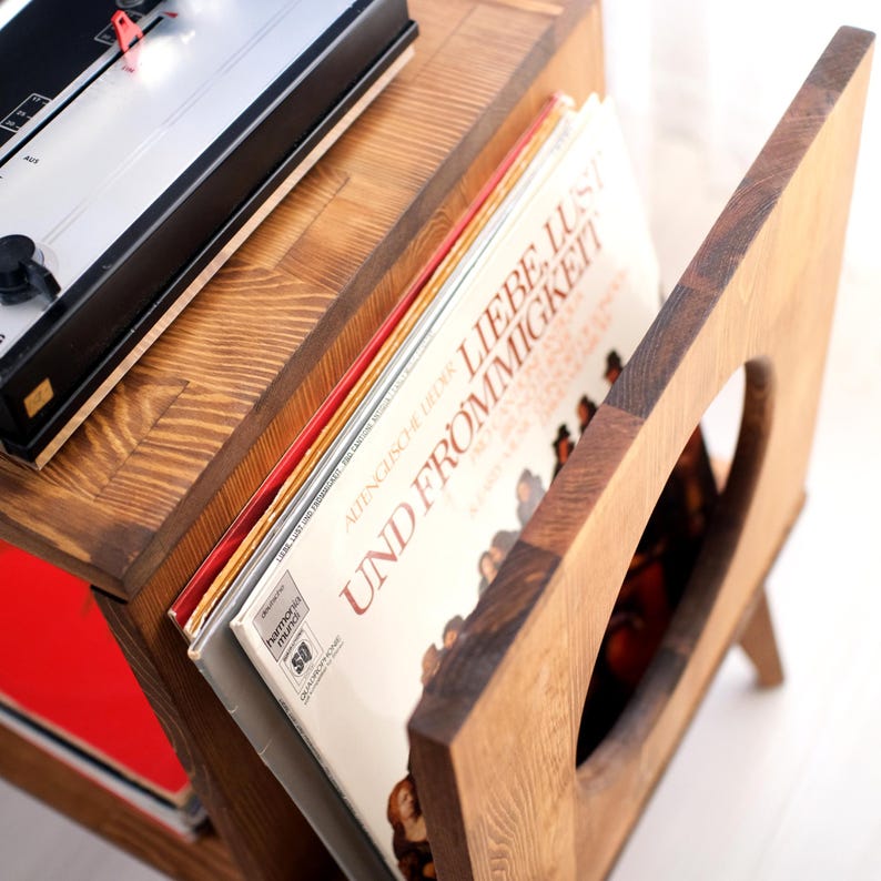 May include: A wooden record player stand with a built-in vinyl record holder. The stand is made of light brown wood and features a cutout design. A vintage record player sits on top, and several vinyl records are visible in the holder.
