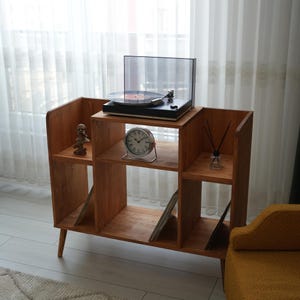 Puede incluir: Un mueble de madera para tocadiscos con tocadiscos, reloj y objetos decorativos. El mueble tiene varios estantes para almacenamiento y exhibición. El tocadiscos es negro con una cubierta transparente. El reloj es plateado con una esfera blanca.