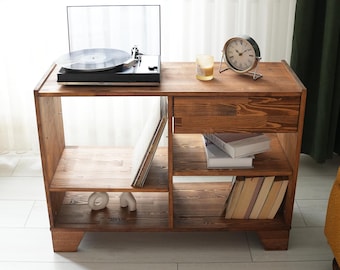 May include: A wooden record player stand with a drawer, shelves, and a turntable on top. The stand is made of dark brown wood and has a rustic look. There are books, records, and decorative items on the shelves.