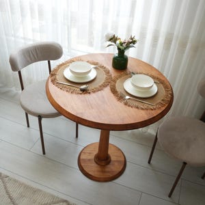 Op de afbeelding: Een ronde houten tafel met een centrale poot en twee beige gestoffeerde stoelen. De tafel is gedekt met twee geweven placemats met franjes, witte kommen op witte borden, zilveren vorken en een kleine groene glazen vaas met kunstbloemen.
