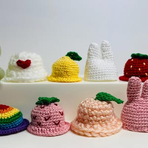 May include: A collection of 10 crocheted hats in various colors and designs, including a strawberry, a lemon, a rainbow, a bunny, and a heart. The hats are all small and would be suitable for dolls or small toys.