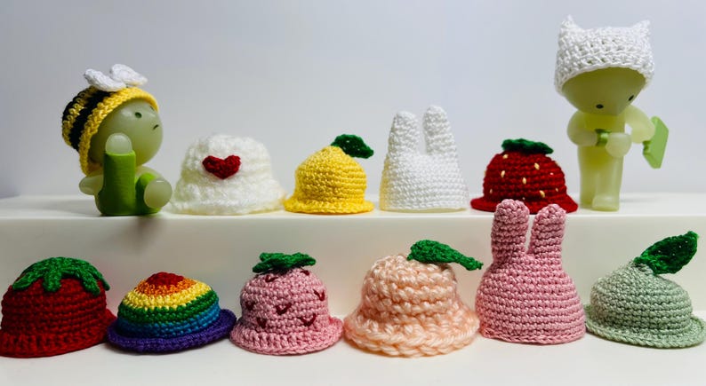 May include: A collection of 10 crocheted hats in various colors and designs, including a strawberry, a lemon, a rainbow, a bunny, and a heart. The hats are all small and would be suitable for dolls or small toys.