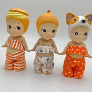 May include: Three dolls with orange and white striped, polka dot, and floral patterned clothing. The dolls have yellow, orange, and white hair and are wearing different hats.