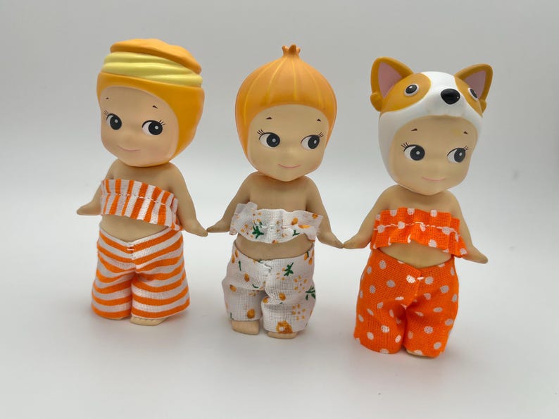 May include: Three dolls with orange and white striped, polka dot, and floral patterned clothing. The dolls have yellow, orange, and white hair and are wearing different hats.