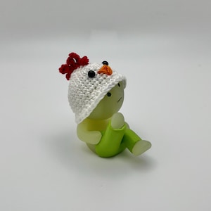 May include: A small, white crocheted chicken hat with a red comb and orange beak sits on top of a green and white figurine. The figurine has a sad expression and is sitting with its legs crossed.