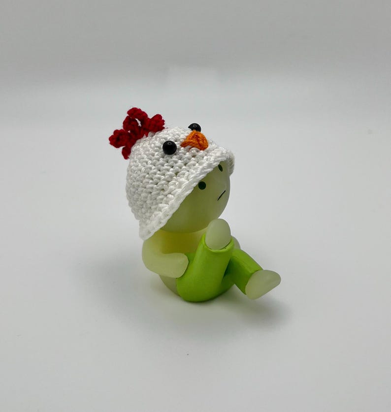 May include: A small, white crocheted chicken hat with a red comb and orange beak sits on top of a green and white figurine. The figurine has a sad expression and is sitting with its legs crossed.
