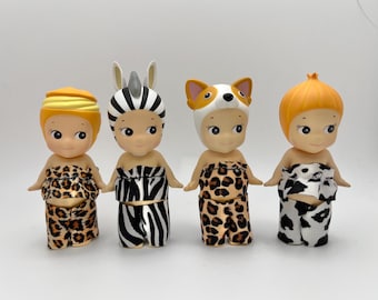 Puede incluir: Cuatro pequeñas figuras con trajes con temática de animales. Una lleva un atuendo con estampado de cebra y un tocado de cebra. Otra tiene un gorro de perro y pantalones con estampado de leopardo. Las otras dos tienen atuendos y tocados con estampado animal.