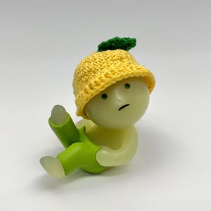 May include: A small, yellow crocheted hat with a green leaf on top. The hat is worn by a small, white figurine with green pants. The figurine has a sad expression on its face.