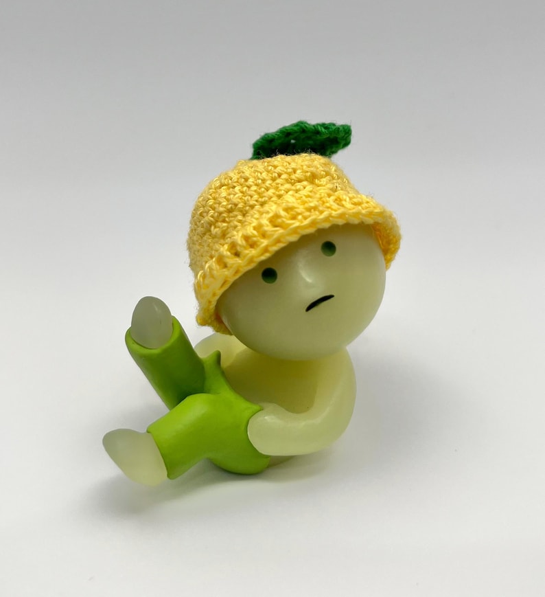 May include: A small, yellow crocheted hat with a green leaf on top. The hat is worn by a small, white figurine with green pants. The figurine has a sad expression on its face.