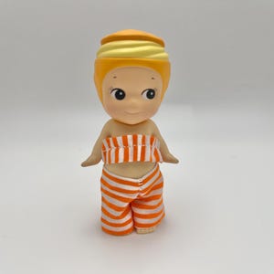 May include: A small, yellow doll with a striped orange and white outfit. The doll has a yellow turban-like hat and a smiling face.