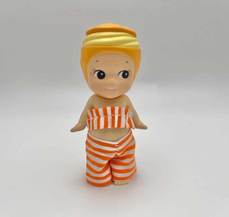 May include: A small, yellow doll with a striped orange and white outfit. The doll has a yellow turban-like hat and a smiling face.