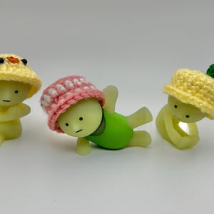 May include: Three small, yellow, glow-in-the-dark figurines wearing crocheted hats. The hats are yellow, pink with white polka dots, and yellow with a green leaf. The figurines are in different poses.