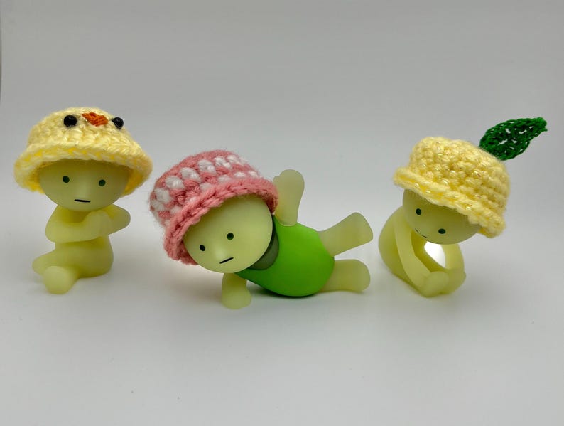 May include: Three small, yellow, glow-in-the-dark figurines wearing crocheted hats. The hats are yellow, pink with white polka dots, and yellow with a green leaf. The figurines are in different poses.