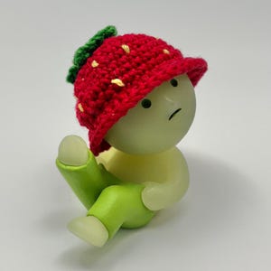 May include: A small, green figurine with a red crocheted strawberry hat. The figurine has a sad expression and is sitting with its legs crossed.