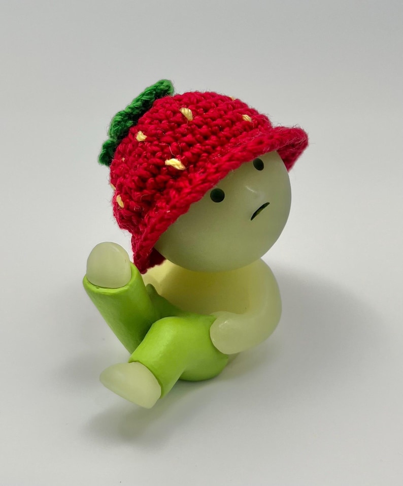 May include: A small, green figurine with a red crocheted strawberry hat. The figurine has a sad expression and is sitting with its legs crossed.