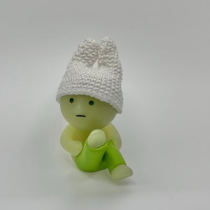 May include: A small, green figurine with a white crocheted hat. The figurine has a round head with a neutral expression and is wearing green pants.