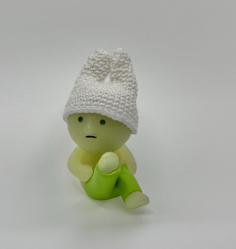 May include: A small, green figurine with a white crocheted hat. The figurine has a round head with a neutral expression and is wearing green pants.