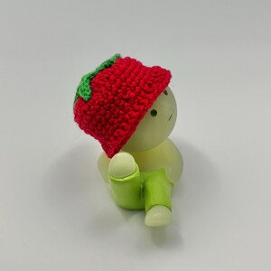 May include: A small, white figurine wearing a red crocheted hat with a green leaf detail and green pants. The figurine is sitting with its legs crossed.